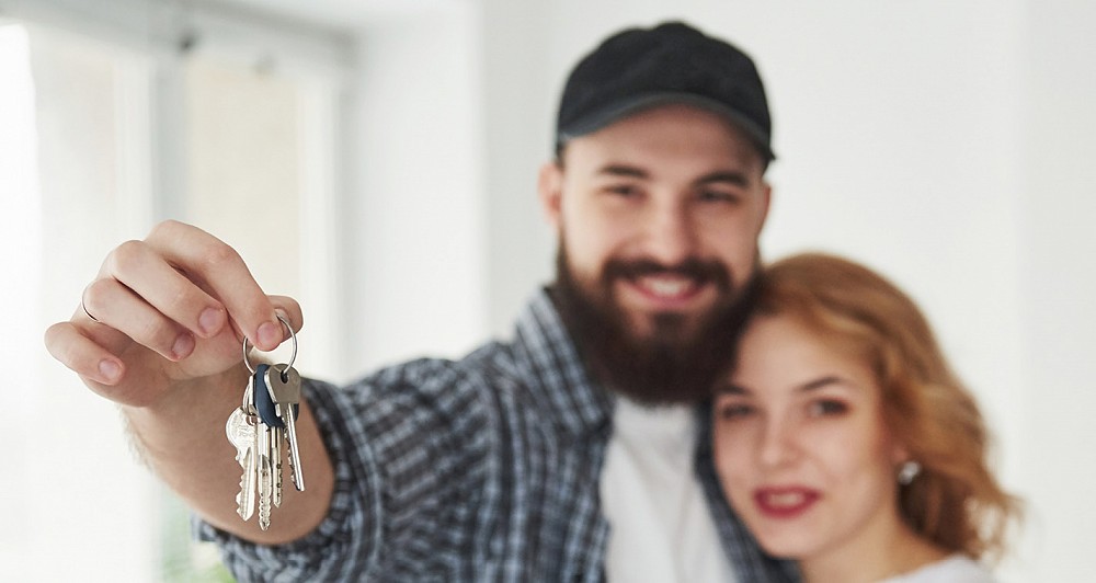 New home owner couple smiling and the man, who has a beard, is holding out the keys to the camera.